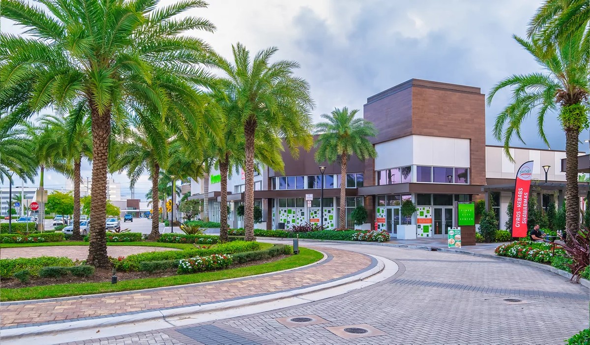 Landscaped entrance to Pines City Center with tall palm trees, flower beds, brick walkways, and storefront signs with gyros, kebabs, and shawarma along Pines Boulevard in Pembroke Pines, Florida