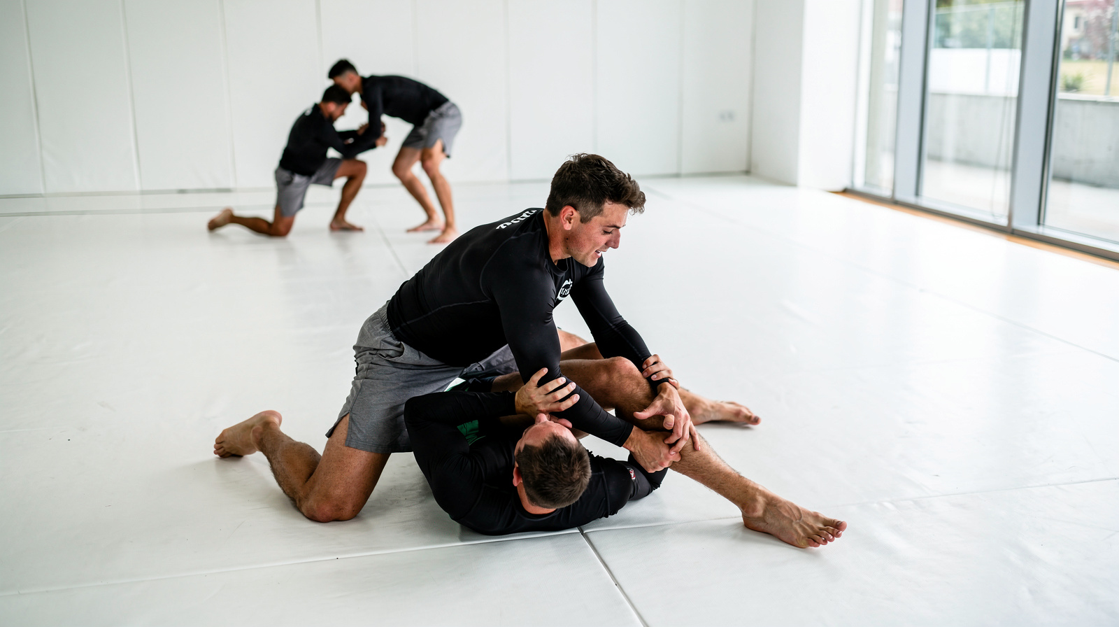 Brazilian jiu-jitsu training session in a modern academy with students in rash guards grappling on white mats