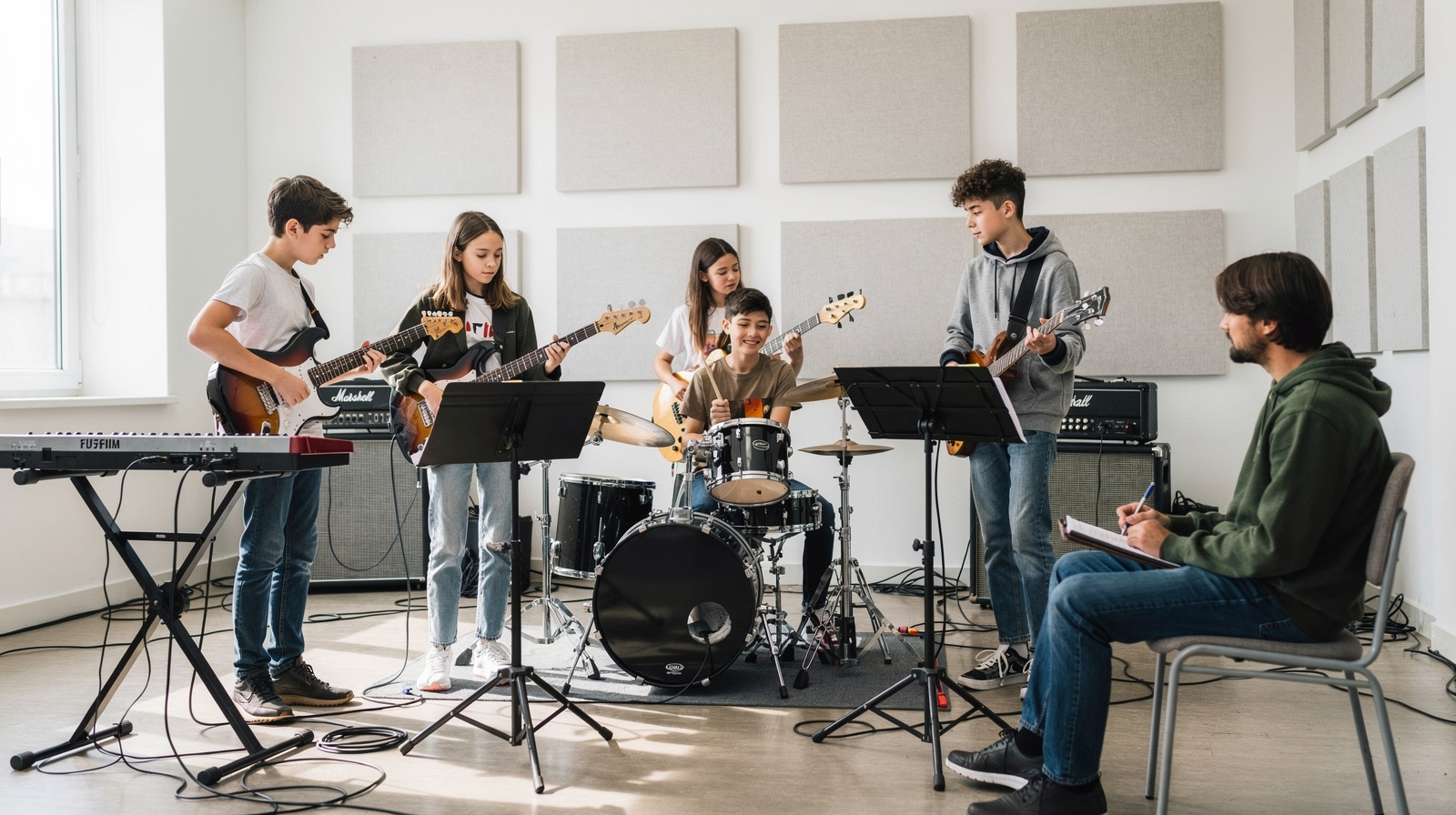 Youth band practice session with teenagers playing electric guitar, bass, drums and keyboard in a bright rehearsal room